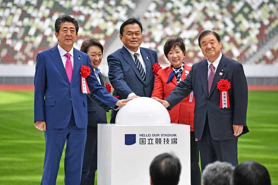 Governor Koike, in a red jacket, stands with Prime Minister Abe and other officials at the ceremony for the completion of the National Stadium
